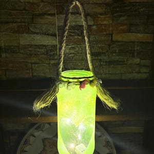 May include: A hanging, glowing green glass jar lantern with a rope handle. The jar is decorated with a floral garland and contains small lights and a heart-shaped design. The lantern hangs against a brick wall.