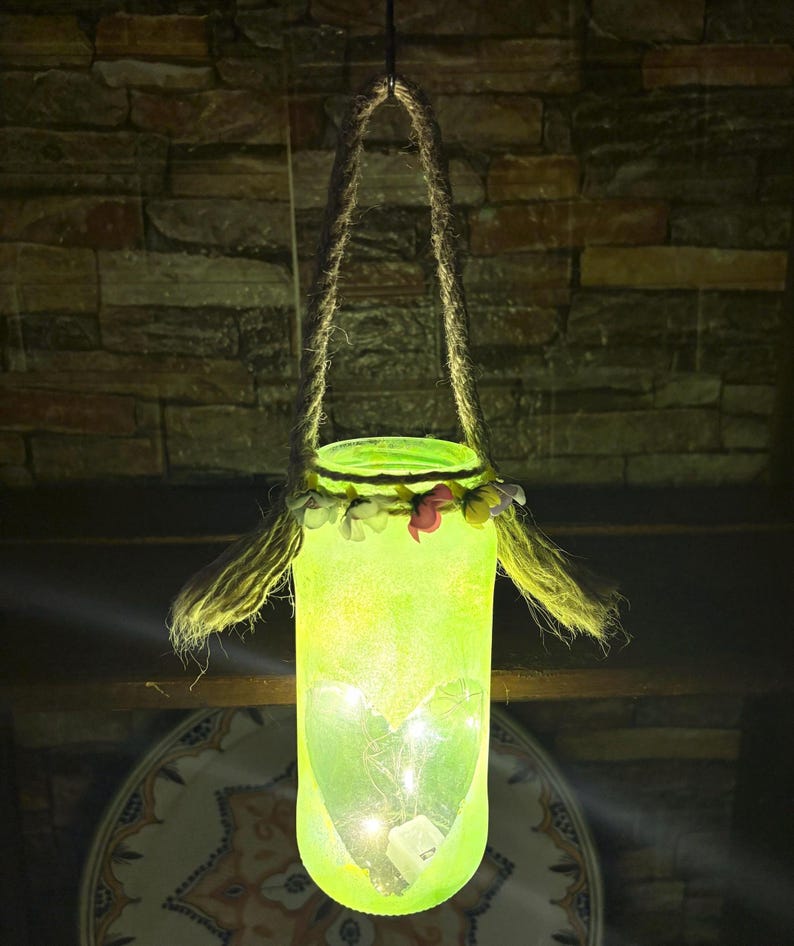 May include: A hanging, glowing green glass jar lantern with a rope handle. The jar is decorated with a floral garland and contains small lights and a heart-shaped design. The lantern hangs against a brick wall.