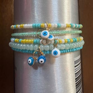 Puede incluir: Conjunto de pulseras de cuentas en tonos turquesa, amarillo, blanco y verde. Pendientes de aro dorados con colgantes de ojo turco azul y blanco cuelgan de las pulseras. Las pulseras están apiladas sobre un objeto cilíndrico plateado.