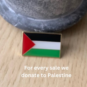 May include: Rectangular pin badge depicting the Palestinian flag, with red, white, black, and green colours, and a gold border. The badge is on a wooden surface. Text on the image reads: "For every sale we donate to Palestine."