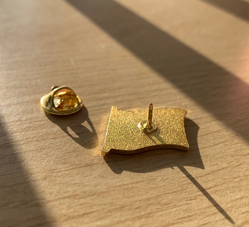 Pu&ograve; includere: Spilla in metallo dorato con un design a bandiera. Il retro della spilla ha una chiusura di sicurezza e uno spillo.