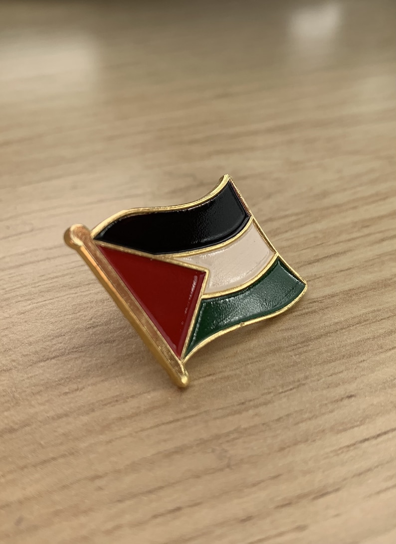 Pu&ograve; includere: Uno spillo dorato con un designo di bandiera palestinese. La bandiera &egrave; nera, bianca, rossa e verde.