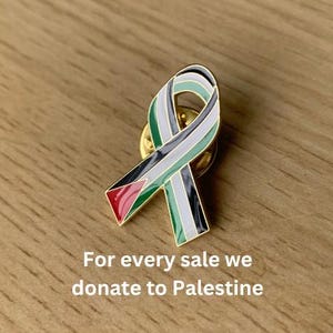 Può includere: Uno spillo dorato a forma di nastro con i colori della bandiera palestinese: nero, bianco, verde e rosso. Lo spillo è su una superficie di legno. Il testo "For every sale we donate to Palestine" è sotto lo spillo.