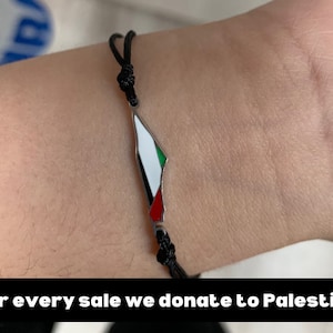 May include: A black string bracelet with a silver charm shaped like the outline of Palestine. The charm is painted with the colors of the Palestinian flag: green, white, red, and black.
