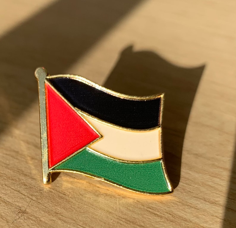 Pu&ograve; includere: Uno spillo dorato con un designo della bandiera palestinese. La bandiera presenta un triangolo rosso, strisce nere, bianche e verdi.