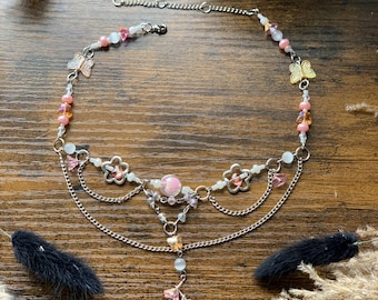 May include: A delicate silver chain necklace with pink and white beads, butterfly charms, and flower accents. The necklace has a layered design with a dangling flower charm.