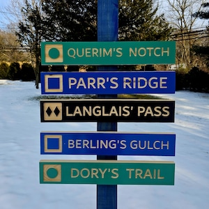 May include: A wooden signpost with five signs pointing in different directions. The signs are painted in different colors, including green, blue, and black. The signs have gold lettering and include the names of trails: "Querim's Notch", "Parr's Ridge", "Langlais' Pass", "Berling's Gulch", and "Dory's Trail".