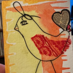 Puede incluir: Tarjeta de arte dibujada a mano con un pájaro amarillo con un ala roja y un corazón dorado. El pájaro está delineado en negro sobre un fondo de acuarela naranja y amarillo. La tarjeta es rectangular y tiene una superficie texturizada.