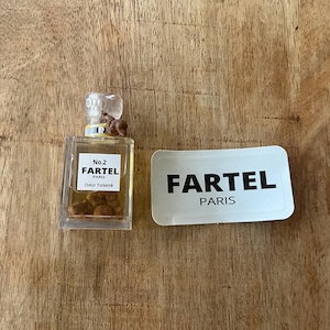 May include: A small, clear perfume bottle with a clear stopper and a white label that reads "No.2 FARTEL PARIS Odor Toilette." Next to it is a white rectangular card with "FARTEL PARIS" printed in black. The items are on a wooden surface.