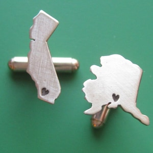 May include: A pair of silver cufflinks shaped like the states of California and Alaska, each with a small heart cutout in the center.