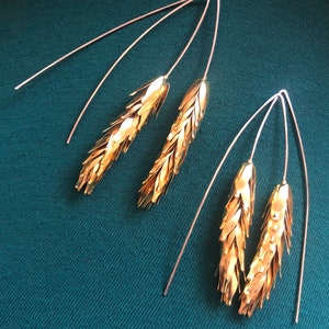 May include: A pair of gold earrings with a wheat design. The earrings are made of thin wire and have a delicate, flowing shape.