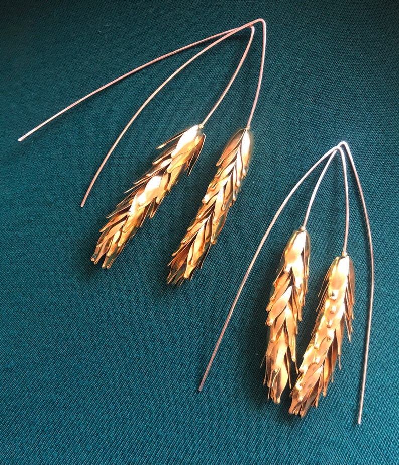 May include: A pair of gold earrings with a wheat design. The earrings are made of thin wire and have a delicate, flowing shape.