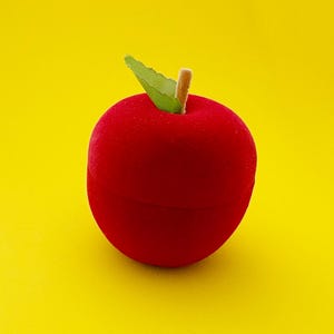 Pode incluir: Uma caixa vermelha em forma de maçã com uma folha verde e um caule castanho. A caixa é feita de um material macio e aveludado. O fundo é amarelo brilhante.
