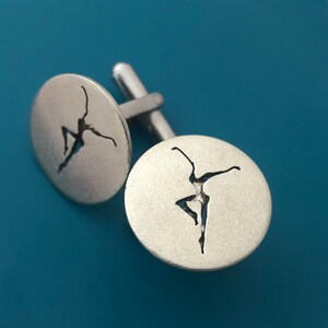 May include: A pair of silver cufflinks with a cutout design of a dancer. The cufflinks are round and have a smooth, polished finish.