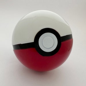 Könnte beinhalten: Ein rotes und weißes Poke Ball Spielzeug mit einem schwarzen Ring in der Mitte und einem kleineren weißen Kreis in der Mitte.