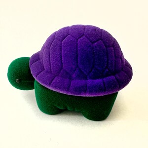 May include: A small purple and green velvet turtle ring box. The turtle has a purple shell with a textured design and a green body.