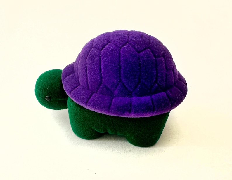 May include: A small purple and green velvet turtle ring box. The turtle has a purple shell with a textured design and a green body.