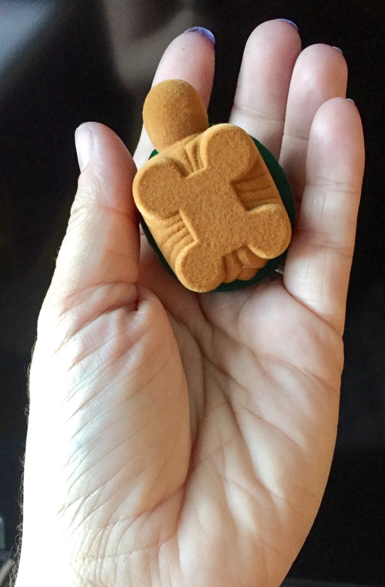 May include: A small, brown velvet ring box with a green velvet lining. The box is shaped like a turtle.