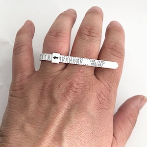 May include: White plastic ring sizer with black markings from size 5 to 17. The sizer is on a person's finger. The sizer has the text "PAT. PEND. 9101693".