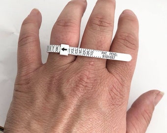 Könnte beinhalten: Ein weißer Kunststoff-Ringgrößenmesser mit schwarzen Markierungen von Größe 5 bis 17. Der Größenmesser befindet sich an einem Finger. Der Größenmesser trägt den Text "PAT. PEND. 9101693".
