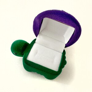 May include: A green turtle-shaped ring box with a purple velvet lid. The box is open and the inside is lined with white velvet.