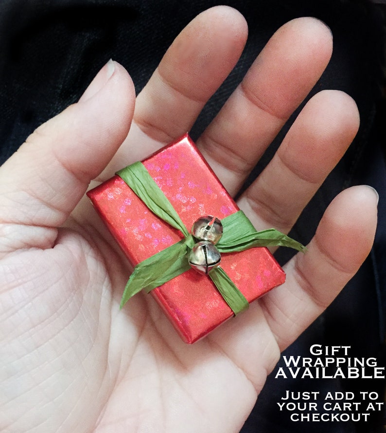 May include: A small red gift box with a green ribbon and silver bells. The box is wrapped in a festive pattern and is held in a hand. The text "Gift Wrapping Available. Just add to your cart at checkout" is visible below the box.