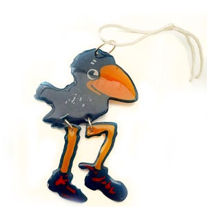 May include: A glossy, dark blue bird ornament with a large orange beak and long, thin orange legs wearing dark blue shoes. The bird has a white eye and is suspended by a thin white string attached to a silver metal jump ring at the top of its head.