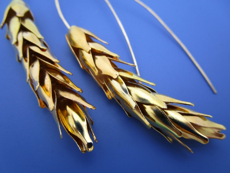 May include: A pair of gold wheat-shaped earrings with silver hooks. The earrings are made of multiple small, overlapping pieces of metal that resemble wheat stalks.