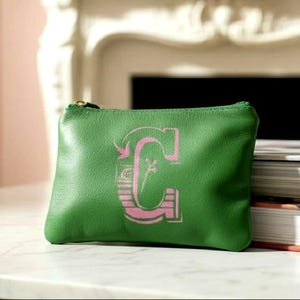 May include: A green leather zippered pouch featuring a large, stylized letter C printed in a light pink hue. The letter C has a decorative, textured design with horizontal lines and a small floral detail integrated into the center of the character.
