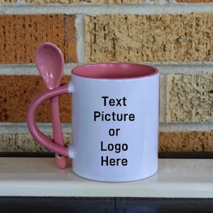 以下が含まれることがあります： ピンクの縁取りと取っ手のある白いセラミック製のマグカップ。マグカップには、取っ手にピンクのスプーンが付いています。マグカップには、「Text Picture or Logo Here」という文字が前面に印刷されています。