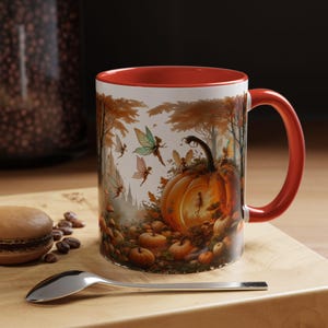May include: A white ceramic mug with a red interior and handle. The mug features a whimsical autumn scene with fairies, pumpkins, and trees. A spoon, a macaron, and coffee beans are on a wooden surface.