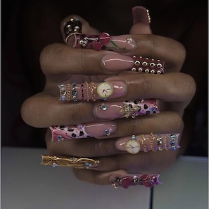 May include: A hand displaying long, decorated acrylic nails. The nails are adorned with gold chains, rhinestones, miniature clocks, and floral and leopard print patterns. The colour scheme includes pink, gold, and burgundy.