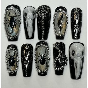 Peut inclure: Un ensemble de dix faux ongles noirs avec divers motifs. Certains ongles pr&eacute;sentent des illustrations d'insectes et d'araign&eacute;es, tandis que d'autres arborent des motifs floraux ou aviaires. Chaque ongle est orn&eacute; d'ornements argent&eacute;s et d'une finition brillante.