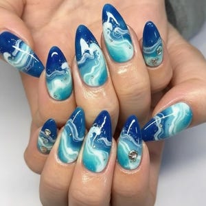 May include: Almond-shaped artificial nails with a blue and white wave design. The nails have a gradient of blue hues, with white swirls and small silver embellishments. The design evokes the look of ocean waves.