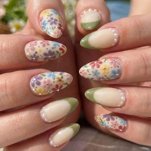 May include: Close-up of manicured nails with a floral design. The nails are painted with a light beige base, adorned with colorful flowers and gold accents. Some nails have a green tip with a pearl border, others are covered in a floral pattern.