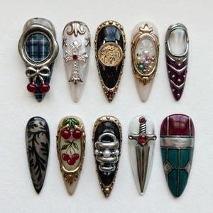 May include: A collection of ten ornate, long, and pointed false nails. Each nail features unique designs, including cherries, crosses, a clock mechanism, pearls, and a sword. The color palette includes silver, gold, red, black, and white.