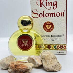 Può includere: Una piccola bottiglia rotonda di vetro di olio di unzione con un tappo dorato, etichettata "King Solomon". La bottiglia è accanto a una scatola con lo stesso nome e un bordo decorativo. L'olio è di 7,4 ml ed è circondato da piccole pietre.