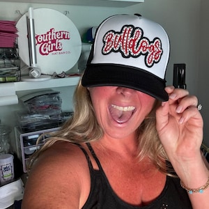 Puede incluir: Una gorra de camionero negra y blanca con un parche blanco que dice "Bulldogs" en rojo con pedrería. La gorra la sostiene una persona con una gran sonrisa.