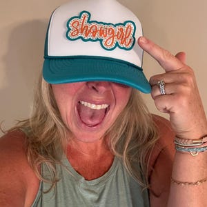 May include: A white trucker hat with a teal brim and a white front panel. The front panel features the word "showgirl" in orange script with an orange outline. The hat is a classic trucker style.