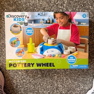 May include: A Discovery Kids motorized pottery wheel kit box featuring a blue and white electric wheel, a lime green tool, paint pots, brushes, and clay pots. The packaging displays text including "Create your own pottery!" and "No baking required!" with an age rating of 8+.