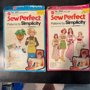 May include: Two rectangular cardboard boxes for Sew Perfect Patterns by Simplicity, numbered 2007 and 2004. The boxes feature illustrations of children, sewing projects, and a sewing machine, with text detailing contents and age requirements.