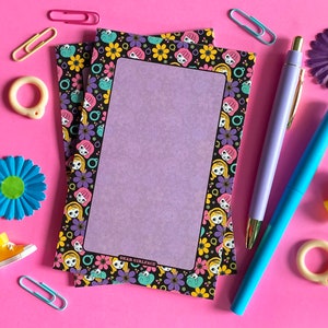 May include: A set of two purple and black patterned notepads with a blank space in the center for writing. The notepads have a border of cartoon faces with flowers and are labeled "Dear Girlface".