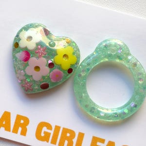 Puede incluir: Un par de pendientes de resina verde con purpurina. Un pendiente tiene forma de corazón con un diseño floral y el otro tiene forma de anillo.