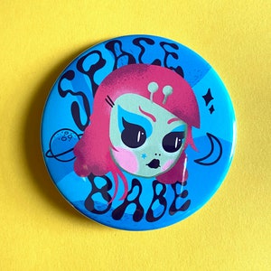 Może przedstawiać: Okrągły guzik w kolorze niebieskim i białym z kreskówkową ilustracją dziewczyny-obcej z różowymi włosami, z półksiężycem i gwiazdami. Tekst "Space Babe" jest napisany stylizowanym fontem wokół dziewczyny-obcej.