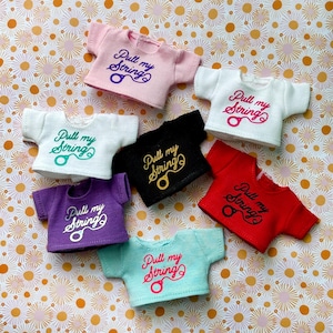 Puede incluir: Siete camisetas en miniatura de diferentes colores con el texto "Pull my String" y un pequeño gráfico de esposas impreso en la parte delantera.