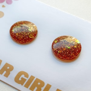 May include: Two round, copper-coloured stud earrings with embedded glitter. The earrings are set against a white background, with the text "AR GIRL" visible in the lower left corner. The glitter reflects gold and green light.