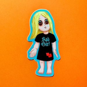May include: A sticker featuring a cartoon girl with blonde hair, red eyes, and a tear. She wears a black t-shirt with the words "Sad Girl" in blue gothic font. The sticker has a blue outline and is set against an orange background.