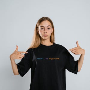 May include: A person wearing a black t-shirt with the text "resist the algorithm" in rainbow colors. The person is pointing to the shirt with both hands.