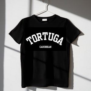 May include: Black t-shirt with the word "TORTUGA" in white arched lettering above the word "CARIBBEAN" in white, displayed on a black rectangular background. The shirt is hanging on a white hanger.