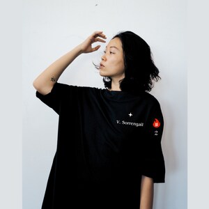May include: A woman wearing a black oversized t-shirt with white text that reads "V. Sorrengail" and a small star above the text. A red flame graphic with the letter "W" inside is on the right sleeve of the shirt.